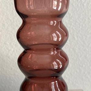 May include: A tall, decorative glass vase in a deep plum color. The vase has a cylindrical top, a series of rounded, bulbous sections, and a clear, rounded base. The glass reflects light, creating highlights and shadows.