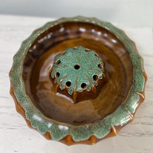 May include: A ceramic flower frog with a brown base and a central, star-shaped holder in a teal-green hue. The rim of the frog has a textured, scalloped edge in the same teal-green color. The frog is designed to hold flowers.