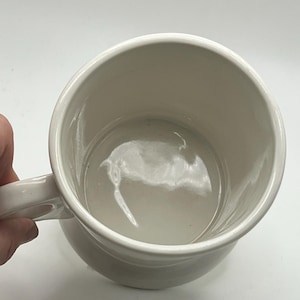 May include: A white ceramic mug with a rounded body and a handle. The interior of the mug is visible, showing a smooth, glossy surface. The mug is set against a plain white background.