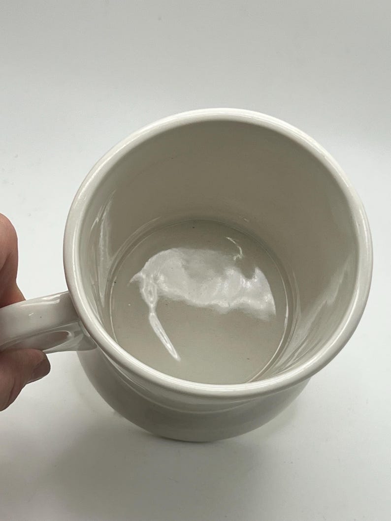 May include: A white ceramic mug with a rounded body and a handle. The interior of the mug is visible, showing a smooth, glossy surface. The mug is set against a plain white background.