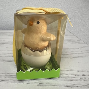 May include: A ceramic chick figurine emerging from a cracked eggshell, presented in a clear plastic box. The chick is light yellow with an orange beak, and the eggshell is white and brown. The box is decorated with a yellow ribbon and sits on a green base.