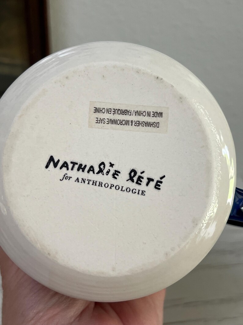 K&ouml;nnte beinhalten: Wei&szlig;er Keramikkrugboden mit dem Text "Nathalie Lete for Anthropologie" in schwarzer Schrift. Der Boden des Krugs hat einen Aufkleber mit der Aufschrift "Dishwasher & Microwave Safe Made in China/Fabriqu&eacute; en Chine."
