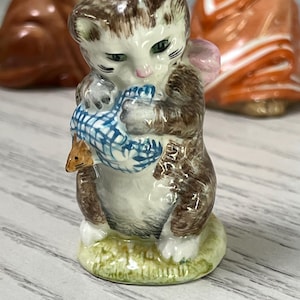 May include: A ceramic figurine of a kitten holding a blue and white patterned bundle, possibly a fish. The kitten has brown and white fur, green eyes, and a pink bow. The figurine is on a green base.
