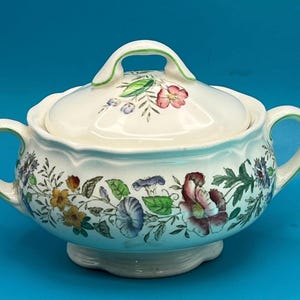 May include: Cream-colored ceramic serving dish with lid, featuring a floral pattern in shades of red, purple, yellow, and green. The dish has two handles and a matching lid with a green-trimmed handle. The floral design wraps around the dish.