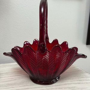 May include: A ruby red glass basket with a handle. The basket has a scalloped rim and a textured, chevron-like pattern. The handle is arched and attached to the sides of the basket. The basket sits on a round base.