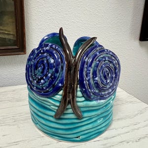 May include: A cylindrical ceramic vessel featuring a turquoise base with horizontal ridged textures. Two prominent dark blue spiral motifs are attached to the front, separated by a central vertical brown element that resembles a butterfly body.