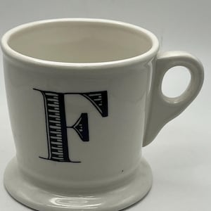May include: A white ceramic mug with a black letter "F" on the front. The mug has a handle and a wide base. The mug is designed for beverages.