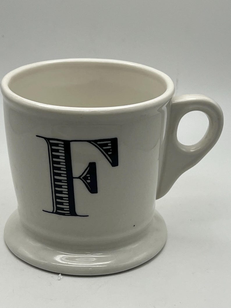 May include: A white ceramic mug with a black letter "F" on the front. The mug has a handle and a wide base. The mug is designed for beverages.