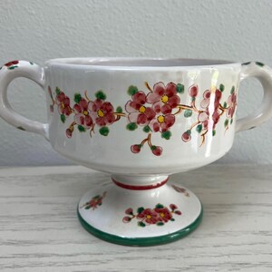 May include: White ceramic pedestal bowl with two handles, adorned with a floral pattern of red flowers and green leaves. The base has a green rim and a red accent. A decorative piece for serving or display.