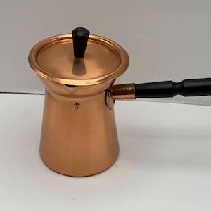 May include: A copper-colored Turkish coffee pot with a black handle and lid. The pot has a rounded base and a narrow spout. The handle is attached to the side of the pot. The lid has a black knob. The number "5" is stamped on the side.