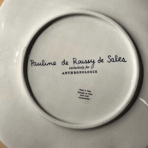 May include: White ceramic plate with a scalloped edge. The underside features the text "Pauline de Roussy de Sales exclusively for ANTHROPOLOGIE." Additional text indicates it was made in China and recommends hand washing.