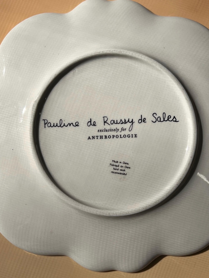 May include: White ceramic plate with a scalloped edge. The underside features the text "Pauline de Roussy de Sales exclusively for ANTHROPOLOGIE." Additional text indicates it was made in China and recommends hand washing.
