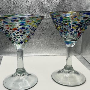 May include: Two clear margarita glasses with colorful, speckled designs. The glasses have a classic triangular bowl shape and a long stem. The speckled design includes red, blue, green, and yellow colors. The glasses are made of glass.