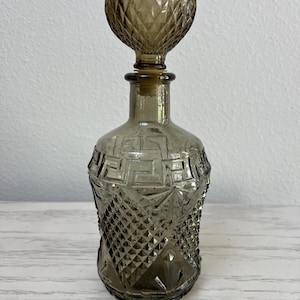 May include: A decorative, smoky-gray glass bottle with a round, textured stopper. The bottle features geometric patterns and a diamond-shaped design. The bottle is approximately 6 inches tall.