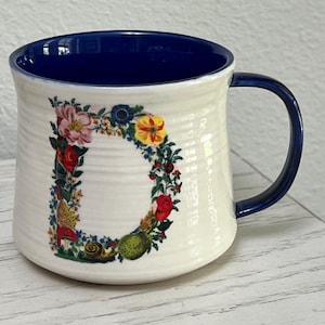 K&ouml;nnte beinhalten: Cremefarbene Keramiktasse mit blauem Innenraum und Henkel. Der Buchstabe "D" ist mit einem Blumenarrangement aus roten, rosa, gelben und blauen Blumen mit gr&uuml;nen Bl&auml;ttern und einer Schnecke verziert.