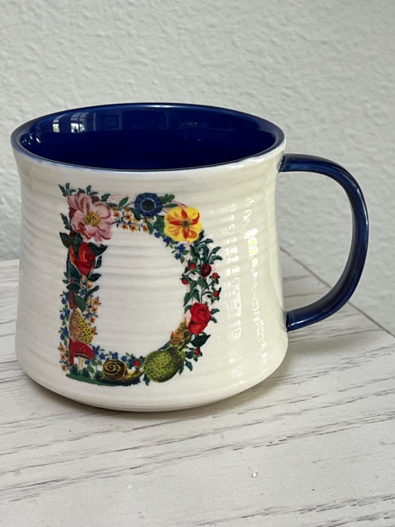 K&ouml;nnte beinhalten: Cremefarbene Keramiktasse mit blauem Innenraum und Henkel. Der Buchstabe "D" ist mit einem Blumenarrangement aus roten, rosa, gelben und blauen Blumen mit gr&uuml;nen Bl&auml;ttern und einer Schnecke verziert.