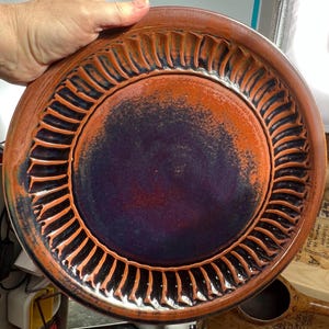May include: A handmade ceramic plate with a unique design. The plate features a dark purple center with an orange and brown rim. The rim has a textured, ridged pattern. The plate is held by a hand.