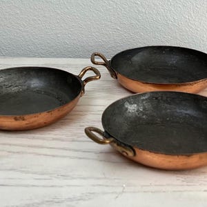 May include: Three small copper pans with dark interiors and loop handles. The pans have a round shape and are arranged on a light-colored surface. The copper has a warm, reddish-brown hue, and the interiors show signs of use.