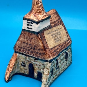May include: A miniature ceramic church with a brown roof and steeple, and a stone-like facade. The text on the side reads "BRITAIN in MINIATURE Handcrafted by TEY POTTERY HAINFORD NORFOLK". The church has small windows and a dark door.