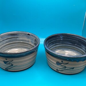 May include: Two ceramic bowls with a blue and gray color scheme. The bowls have a dark blue rim and a design on the side. The bowls are handmade and have a unique, artistic look. The bowls are perfect for serving food or as decorative pieces.