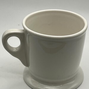 May include: A cream-colored ceramic mug with a rounded handle and a circular base. The mug has a simple, classic design, suitable for coffee or tea. The mug is empty and sits on a white surface.
