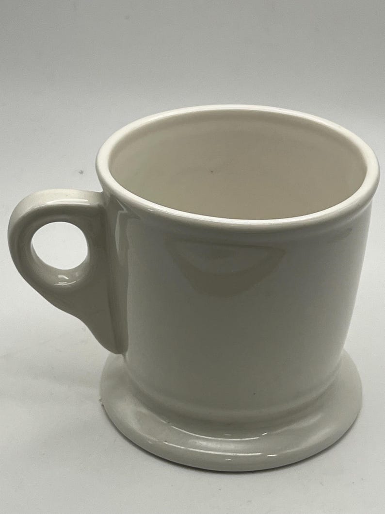 May include: A cream-colored ceramic mug with a rounded handle and a circular base. The mug has a simple, classic design, suitable for coffee or tea. The mug is empty and sits on a white surface.