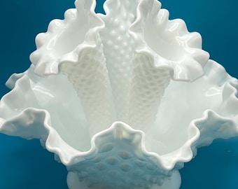 May include: A white glass bowl with a ruffled, scalloped edge and a textured, hobnail pattern. The bowl has a wide, open design and sits on a round base. The background is a solid blue.