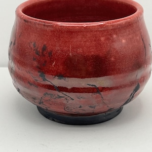 May include: A handmade, red ceramic bowl with a dark base. The bowl has a rounded shape with a wide opening and a textured surface. Black markings add detail to the red glaze. This unique bowl is perfect for serving or display.