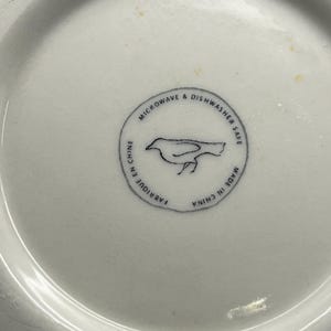 May include: A white ceramic dish with a circular design. The design includes a bird illustration and the text "MICROWAVE & DISHWASHER SAFE", "FABRIQUE EN CHINE", and "MADE IN CHINA".