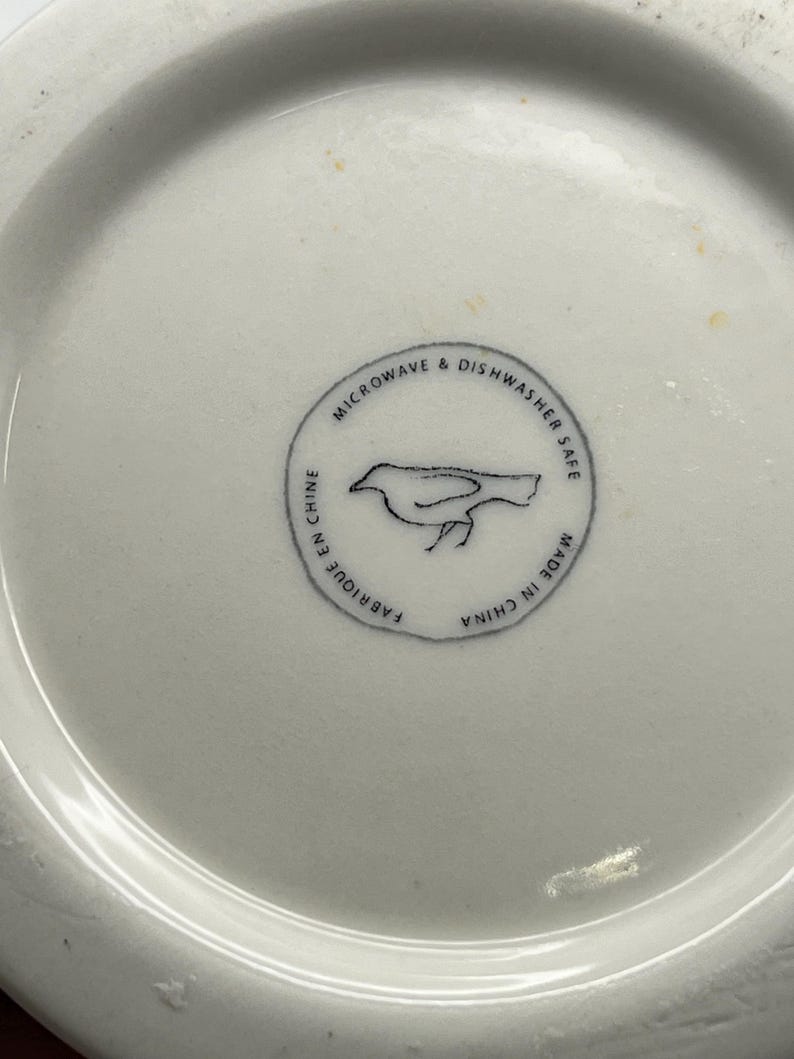 May include: A white ceramic dish with a circular design. The design includes a bird illustration and the text "MICROWAVE & DISHWASHER SAFE", "FABRIQUE EN CHINE", and "MADE IN CHINA".