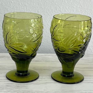 May include: Two green-tinted glass goblets. The glasses have a floral design etched into the surface. The base and lower portion of the glasses are a darker shade of green. The goblets are on a light-colored surface.