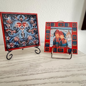 May include: Two decorative square tiles on black metal stands. One tile features a floral design in blue, red, and white against a black background. The other tile depicts two people in traditional clothing, with a snowy landscape.