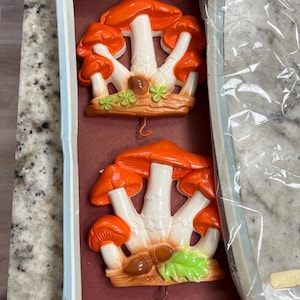 May include: Two ceramic mushroom wall decorations. Each features orange caps, white stems, and a base with acorns and green leaves. The mushrooms are displayed in a box with a red interior.