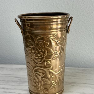 May include: A gold-colored metal vase with a floral pattern. The vase has two small handles and a cylindrical shape. The floral design is embossed on the surface of the vase.
