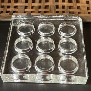 May include: A clear glass square with nine circular indentations. The glass has a textured, uneven edge. The background features a woven wooden panel and a dark surface.