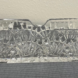 May include: A clear, rectangular glass dish with two handles. The dish has a cut-glass design with a series of diamond and fan-shaped patterns. The dish is sitting on a gray surface.