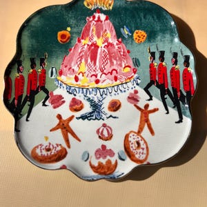 May include: A decorative ceramic plate with a scalloped edge. The plate features a whimsical design with a red and pink cake, gingerbread men, donuts, and marching soldiers in red coats and black hats.