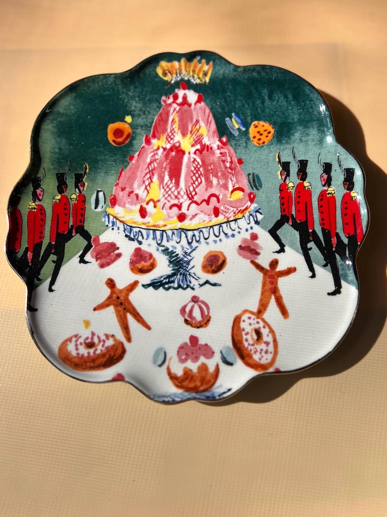 May include: A decorative ceramic plate with a scalloped edge. The plate features a whimsical design with a red and pink cake, gingerbread men, donuts, and marching soldiers in red coats and black hats.