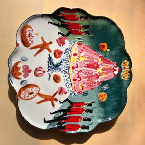 May include: A decorative ceramic plate with a scalloped edge, featuring a whimsical design. The plate is divided into two sections: one with baked goods and the other with a red and green design. The plate has a unique, artistic style.