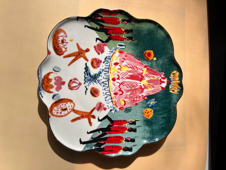 May include: A decorative ceramic plate with a scalloped edge, featuring a whimsical design. The plate is divided into two sections: one with baked goods and the other with a red and green design. The plate has a unique, artistic style.