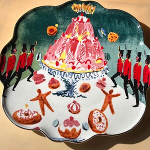 May include: A decorative ceramic plate with a scalloped edge. The plate features a whimsical design with a large pink cake, gingerbread men, donuts, and marching soldiers in red coats. The background is green and white.