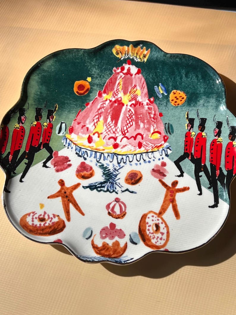 May include: A decorative ceramic plate with a scalloped edge. The plate features a whimsical design with a large pink cake, gingerbread men, donuts, and marching soldiers in red coats. The background is green and white.