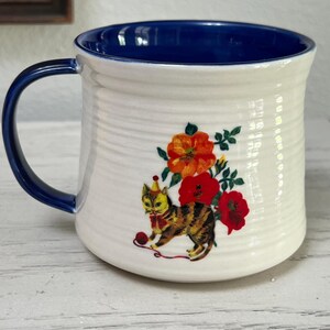 K&ouml;nnte beinhalten: Wei&szlig;er Keramikbecher mit blauem Innenraum und Henkel. Der Becher zeigt ein skurriles Design einer Katze mit Partyhut, roter Fliege und Wollkn&auml;uel, umgeben von orangefarbenen und roten Blumen.