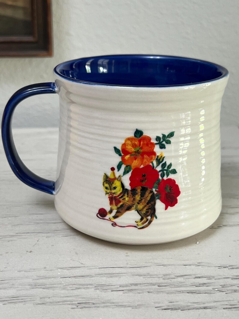 K&ouml;nnte beinhalten: Wei&szlig;er Keramikbecher mit blauem Innenraum und Henkel. Der Becher zeigt ein skurriles Design einer Katze mit Partyhut, roter Fliege und Wollkn&auml;uel, umgeben von orangefarbenen und roten Blumen.