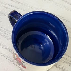 K&ouml;nnte beinhalten: Eine wei&szlig;e Keramiktasse mit blauem Innenraum und Henkel. Die Au&szlig;enseite hat ein Blumendesign mit roten, orangefarbenen und gr&uuml;nen Akzenten. Die Tasse ist leer und steht auf einer hellen Oberfl&auml;che.