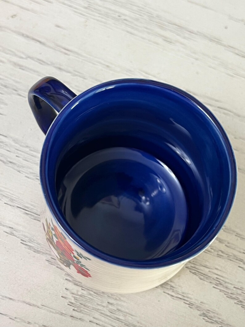 K&ouml;nnte beinhalten: Eine wei&szlig;e Keramiktasse mit blauem Innenraum und Henkel. Die Au&szlig;enseite hat ein Blumendesign mit roten, orangefarbenen und gr&uuml;nen Akzenten. Die Tasse ist leer und steht auf einer hellen Oberfl&auml;che.