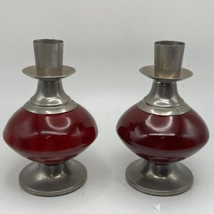 May include: Two matching candle holders. Each has a rounded, deep red glass base, a silver-colored metal stem, and a silver-colored metal cup to hold a candle. The candle holders are on a white background.