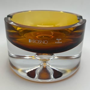May include: A round, amber-colored glass ashtray with a clear base. The ashtray has a thick, rounded rim and a shallow bowl. The base is clear and has a pedestal-like design. The text "KROSNO" and "Made in Poland" are visible.