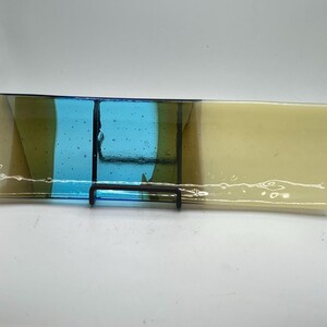May include: A rectangular glass dish with a curved shape, featuring sections of light yellow, blue, and olive green. The glass is supported by a black metal stand. The dish is likely used for decorative purposes.