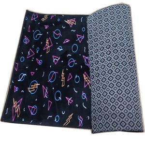 K&ouml;nnte beinhalten: Ein schwarzer rechteckiger Teppich mit einem Retro-Design der 80er Jahre. Der Teppich zeigt neonrosa, blaue und gelbe geometrische Formen, darunter Blitze, Kreise und Dreiecke. Der aufgerollte Abschnitt zeigt ein grau-wei&szlig;es Rautenmuster.